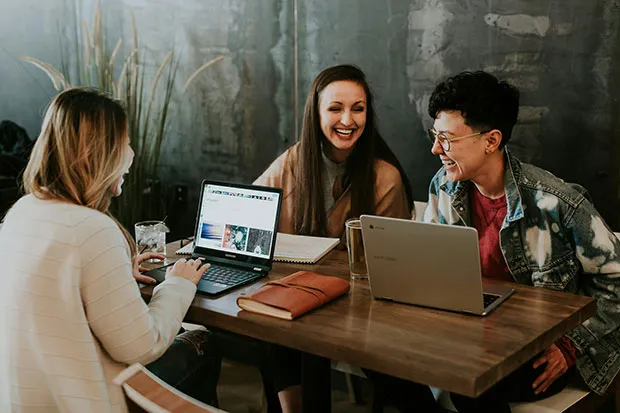 Three students studying and laughing with laptops at a coffee shop