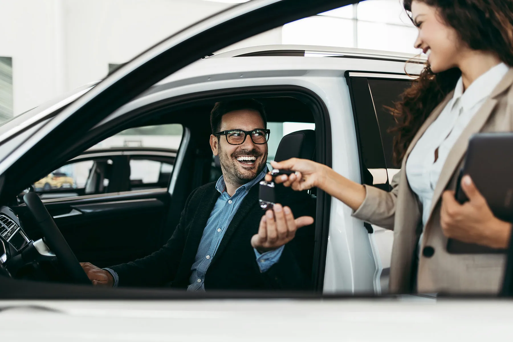 A smiling man sitting in a new car receives his keys from the saleswoman